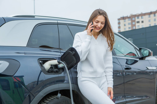 ガソリンスタンドで電気自動車を充電する若い女性は,スマートフォンを使用しています. エコ燃料コンセプト。 環境に優しい輸送の概念. 充電ステーションからのバッテリーの充電. - 写真・画像
