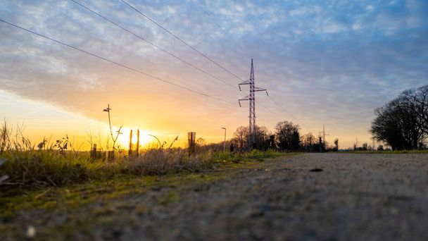 This image presents a ground-level view of a country road at sunset, with the focus on the power lines against the warm glow of the setting sun. The low angle emphasizes the rough texture of the road - Photo, Image