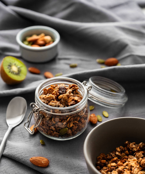 Zelfgemaakte granola in een pot en kom op een donkere linnen ondergrond met noten en fruit. Het concept van gezonde en voedzame volkorenmuesli als ontbijt.  - Foto, afbeelding