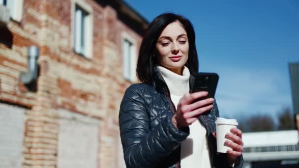 Portrait of brunette woman using electronic device while standing with plastic coffee cup on blurred background. Smiling lady using food delivery service for getting shipment during walk outside. - Footage, Video