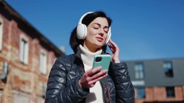 Rozptýlená žena v teplých šatech poslouchá pop song přes bezdrátová sluchátka s telefonem v ruce. Sebevědomá žena používající přenosná zařízení při pilném zpívání textů s hitem. - Záběry, video
