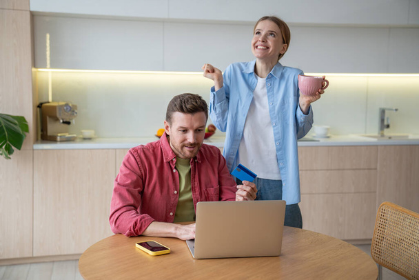 Happy excited married couple rejoices in kitchen after receiving message on laptop with amazing offer, good news. Dream of owning home has become reality thanks to attractive offer for young family. - Photo, Image