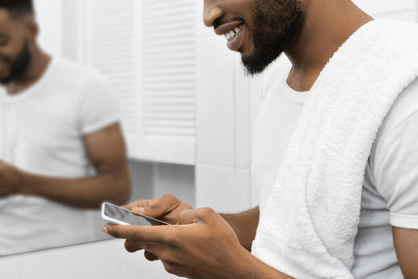 Mosolygó afro ember törülközővel olvasás üzenet okostelefon a fürdőszobában. Férfi fürdőszobai koncepció - Fotó, kép