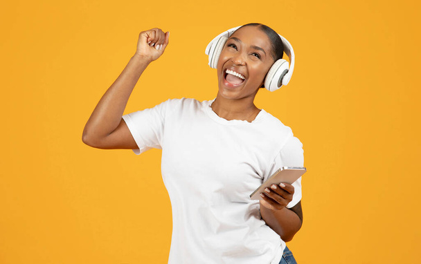 Milenial mujer negra en auriculares inalámbricos con teléfono inteligente disfruta de melodías en el fondo del estudio amarillo, bailando con música y divirtiéndose. Concepto de tecnología moderna y ocio - Foto, Imagen