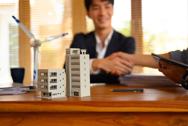 Gros plan du modèle de construction à côté du modèle d'éolienne sur la table de réunion contre les mains de la cabane des gens d'affaires. - Photo, image