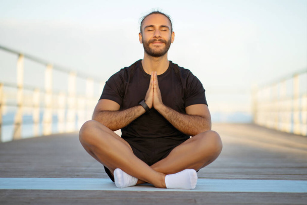 Friedlicher junger Fitness-Typ meditiert mit geschlossenen Augen am Seebrücke, klammert die Hände zusammen und sucht Entspannung und Gleichgewicht in seiner Zen-artigen Meditation im Freien, genießt morgens Yoga-Praxis - Foto, Bild