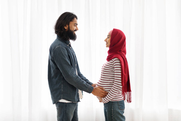 Junges religiöses muslimisches Paar erwartet Baby, steht zusammen am Fenster zu Hause, fürsorglicher Millennial-arabischer Ehemann berührt zärtlich Bauch seiner schwangeren Frau im Hijab, Seitenansicht mit Kopierraum - Foto, Bild