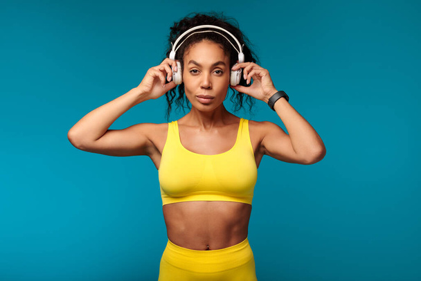 Fitness vibe. Motivated African American woman in yellow activewear stands confidently in studio, putting on wireless earphones, ready for healthy workout while listening to music on blue backdrop - Photo, Image