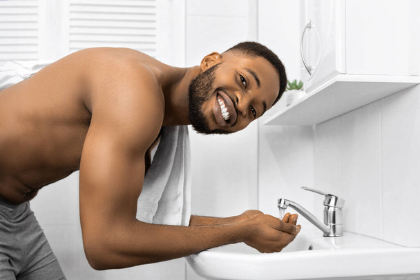 Çıplak kaslı gövde banyoda yüz yıkama ile gülümseyen afro adam, kamera sabah hijyen konsepti bakarak, kopya alanı - Fotoğraf, Görsel