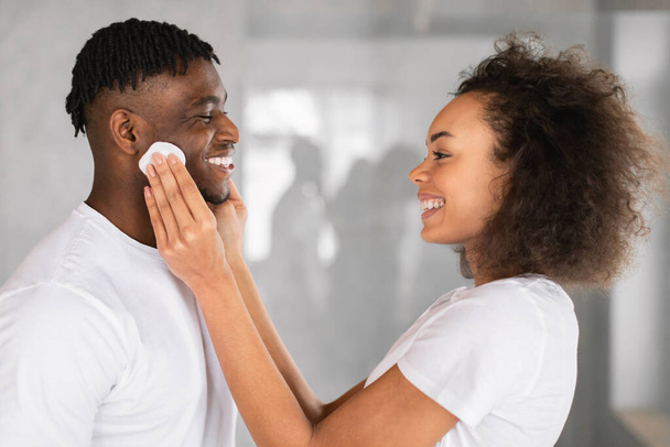 Skincare routine. Loving black couple enjoys domestic skin care moment together, as wife cleansing husbands facial skin with cotton pads in modern bathroom, side view shot - Photo, Image