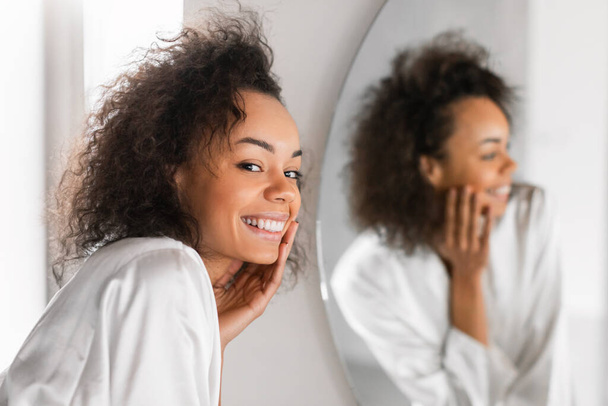 Die attraktive junge Afroamerikanerin mit weichem, feuchtigkeitsspendenden Teint berührt ihr Gesicht während der täglichen Hautpflege im Badezimmer und posiert in einem seidenen Bademantel neben einem Spiegel. Selektiver Fokus - Foto, Bild