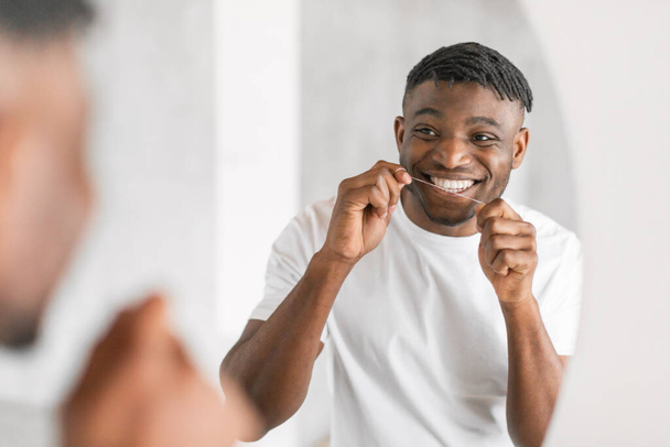Uśmiechnięty młody czarny mężczyzna nici dentystycznej zębów w pobliżu lustra, gdy stoi w łazience, podkreślając swoje zaangażowanie w opiekę jamy ustnej i higieny jamy ustnej, czyszczenie zębów z nici rano - Zdjęcie, obraz