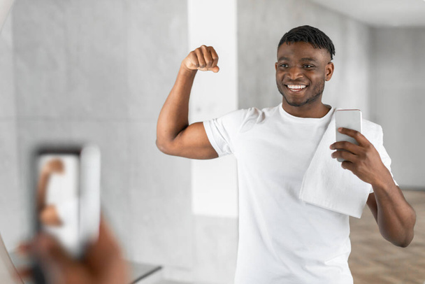 Hymyilevä lihaksikas musta kaveri ottaa selfie käyttäen älypuhelinta kylpyhuoneessa, osoittaa hauikset katsomalla peilikuvausta, valmistautumalla aamulla ja jakamalla hemmottelua sosiaalisessa mediassa - Valokuva, kuva
