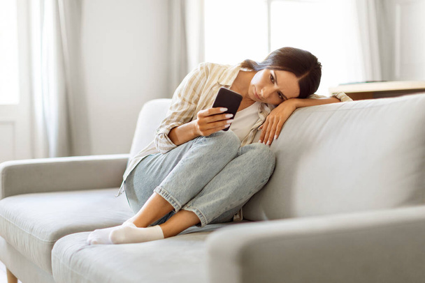 Warten auf seinen Anruf. Aufgebrachte junge Frau schaut auf Smartphone-Bildschirm, während sie zu Hause auf der Couch sitzt, depressive Europäerin hält Handy, liest SMS oder Online-Nachrichten, kopiert Raum - Foto, Bild