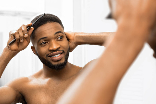 Улыбающийся афро-мужчина с обнаженным туловищем расчесывает волосы и смотрит в зеркало в ванной комнате
. - Фото, изображение