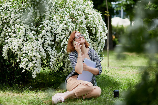 Zonnige werkdag geniet van een freelancer geniet van een gesprek in een bloemenpark. Roodharige vrouw spreekt per telefoon met laptop. - Foto, afbeelding