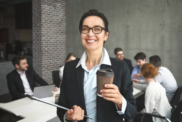 Glückliche Geschäftsfrau mit Kaffee zum Mitnehmen im Büro, posiert bei Vorstandssitzung gegen Kollegen - Foto, Bild