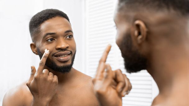 Улыбающийся афро-человек наносит крем на лицо в ванной, отражаясь в зеркале. Человек в ванной концепции - Фото, изображение
