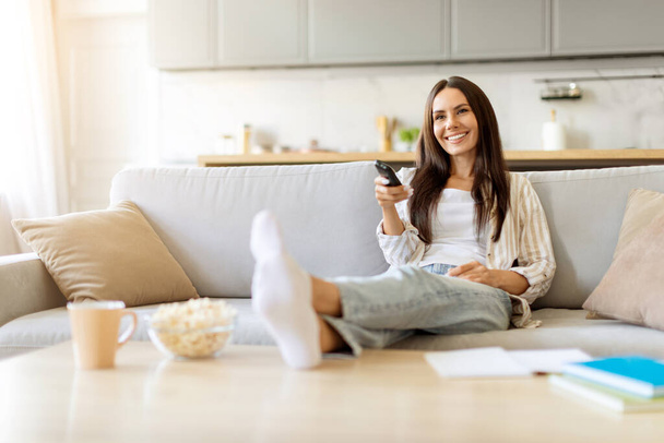 Улыбающаяся молодая женщина, лежащая на диване с поднятыми ногами, держащая пульт от телевизора, счастливая миллениальная леди, отдыхающая на диване с чашей попкорна поблизости, смотрящая телевизор и расслабляясь в уютной гостиной дома - Фото, изображение