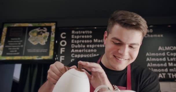 エプロンの男性バリスタは,現代のカフェでコーヒーを注ぐ準備しながら,ケトルからカップに水を充填します. 専門の喫茶店でラテアートを作るバリスタ. コーヒーを作る. - 映像、動画