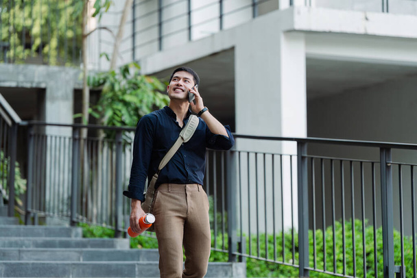 Usmívající se asijský podnikatel držet opakovaně použitelný ekologický pohár a pomocí mobilu, zatímco na cestě domů v parku. - Fotografie, Obrázek