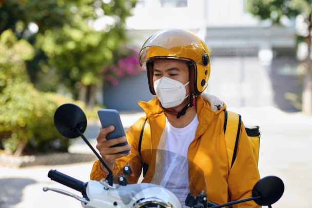 Hombre de entrega seria sentado en moto al leer la próxima notificación de pedido en el teléfono inteligente - Foto, imagen