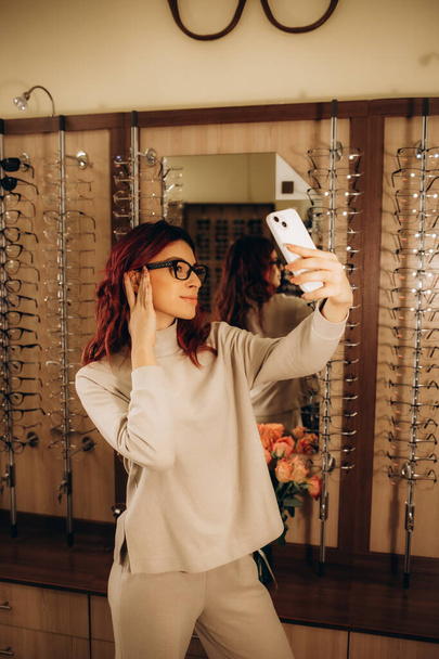 Arckép a vonzó nő a vásárlás álló optikus boltban, hogy szelfi divatos szemüveg és mosolygós. Kiváló minőségű fénykép - Fotó, kép