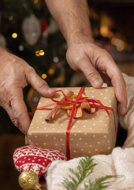 Mikulás kezét fel egy ajándék díszített szárított gyümölcsök közelében hangulatos szív és pulóver ellen karácsonyfa fények, közelről. Téli ünnepek hangulata a nagymamák kezét szóló ajándék doboz  - Fotó, kép
