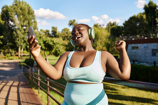 Μια Αφροαμερικανή γυναίκα, με σώμα θετικό και καμπυλωτό, που φοράει ακουστικά ενώ κρατάει ένα κινητό έξω.. - Φωτογραφία, εικόνα