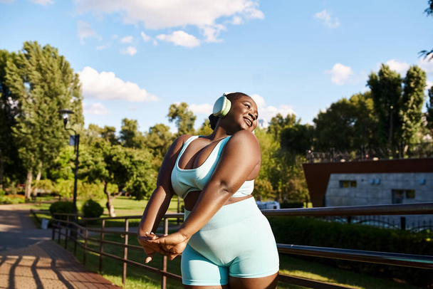 Een Afro-Amerikaanse vrouw met een golvend lichaam, draagt een blauwe sportbeha en korte broek, luisterend naar muziek buiten. - Foto, afbeelding