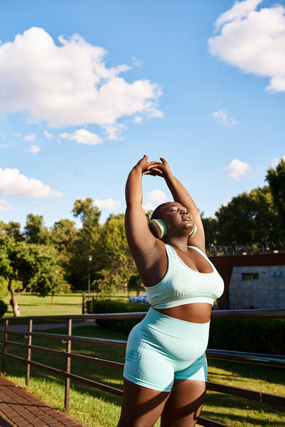 青いトップとショートパンツのアフリカ系アメリカ人女性がエネルギー的に彼女の腕を屋外に伸ばし,体のポジティブさを体現しています.. - 写真・画像