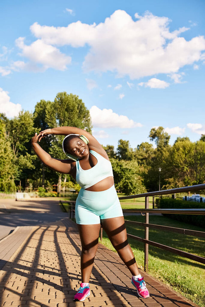 An African American woman in blue shorts and a white tank top stretches her arms outdoors - Photo, Image