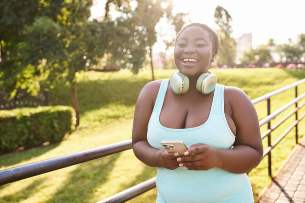 Uma mulher afro-americana confiante, usando fones de ouvido, gosta de música enquanto segura um telefone celular ao ar livre. - Foto, Imagem