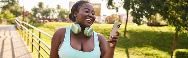 Uma mulher afro-americana ouve alegremente música em seu celular enquanto usa fones de ouvido ao ar livre. - Foto, Imagem