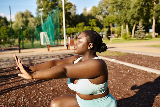 Una mujer afroamericana con un sujetador deportivo azul estira los brazos al aire libre, abrazando su positividad corporal y su viaje de fitness. - Foto, imagen
