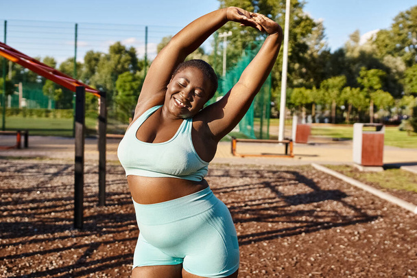 Una donna afroamericana dal corpo sinuoso, in reggiseno sportivo blu, che allunga le braccia all'aperto. - Foto, immagini