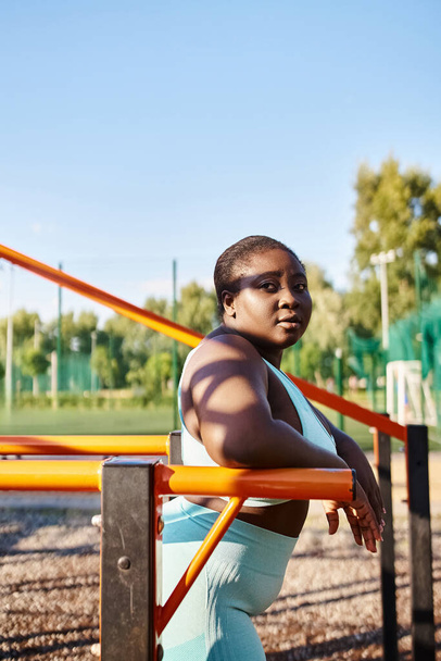 Egy afro-amerikai nő sportruházatban, testpozitivitással, egy padon ül a parkban, körülvéve természettel.. - Fotó, kép