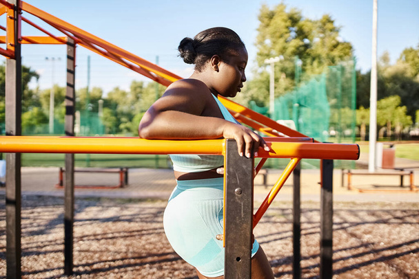 スポーツウェアのアフリカ系アメリカ人女性は,屋外の遊び場のレールにエレガントに傾いている間,体のポジティブさを示しています. - 写真・画像