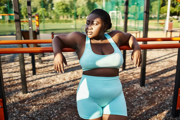 Μια Αφρο-Αμερικανίδα γυναίκα στα αθλητικά στέκεται ευτυχισμένη μπροστά σε μια παιδική χαρά, ενσαρκώνοντας τη θετικότητα και την ενδυνάμωση του σώματος.. - Φωτογραφία, εικόνα