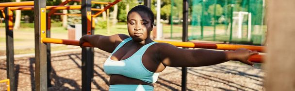 Μια Αφροαμερικανή γυναίκα με αθλητικά να στέκεται μπροστά σε μια παιδική χαρά, επιδεικνύοντας θετικότητα και δύναμη στο σώμα.. - Φωτογραφία, εικόνα