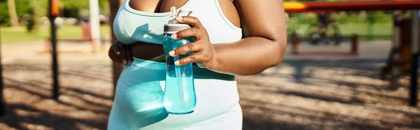 Mujer afroamericana curvilínea en ropa deportiva sosteniendo una botella, rodeada de naturaleza, encarnando la positividad corporal mientras hace ejercicio en un parque. - Foto, Imagen