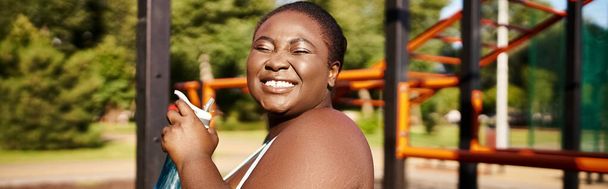 Una mujer afroamericana en ropa deportiva se para con confianza frente a un patio de recreo, encarnando la positividad y la fuerza del cuerpo. - Foto, Imagen