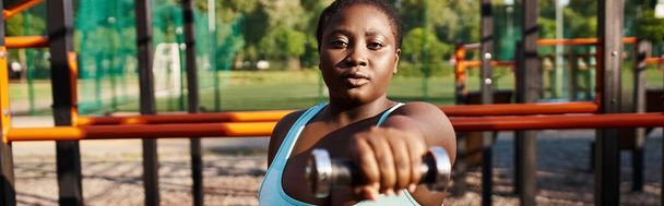Zakřivená Afroameričanka v modrém sportovním oblečení sebejistě drží činku v rukou, vyzařuje z ní půvab a síla. - Fotografie, Obrázek