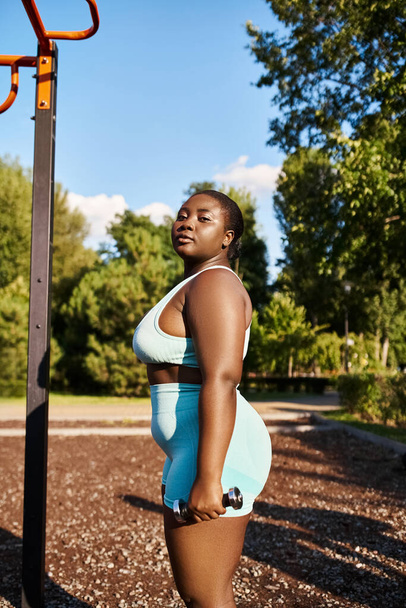 Eine kurvige Afroamerikanerin in Sportbekleidung steht neben einer Stange in einem Park. - Foto, Bild