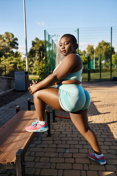 Afrikkalainen amerikkalainen nainen urheiluvaatteissa treenaa sulavasti puisella penkillä luonnon ympäröimänä, sisältäen kehon positiivisuuden.. - Valokuva, kuva
