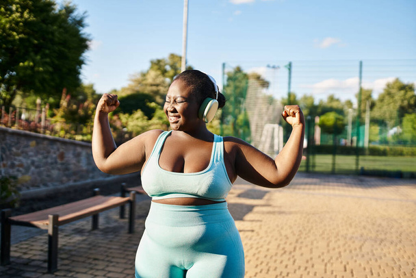 An African American woman in a sports bra and leggings flexes her muscles confidently outdoors. - Photo, Image