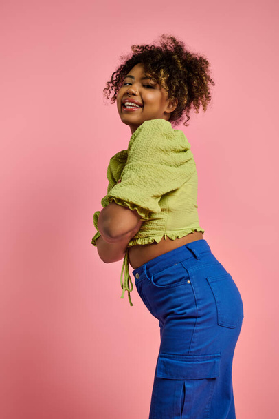 スタイリッシュなアフリカ系アメリカ人女性は活気に満ちた黄色のトップとブルーのズボンでポーズします. - 写真・画像