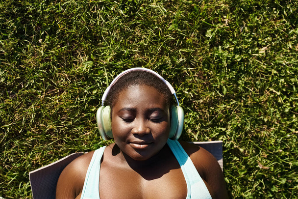 Afro-Amerikalı bir kadın spor kıyafetleriyle doğada dinleniyor, otların üzerinde yatarken kulaklıkla müzik dinliyor.. - Fotoğraf, Görsel