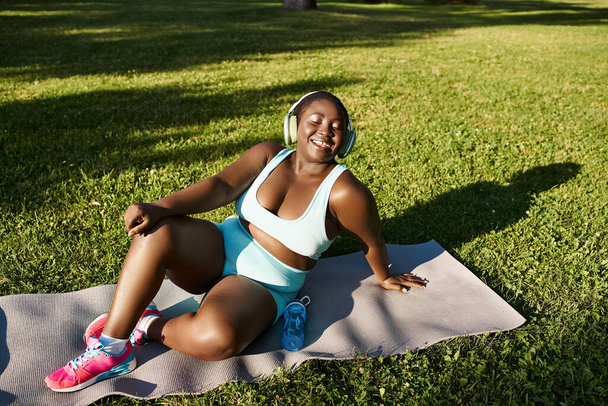 Een bochtige Afro-Amerikaanse vrouw in sportkleding zit sierlijk op een yoga mat beoefenen mindfulness buiten. - Foto, afbeelding
