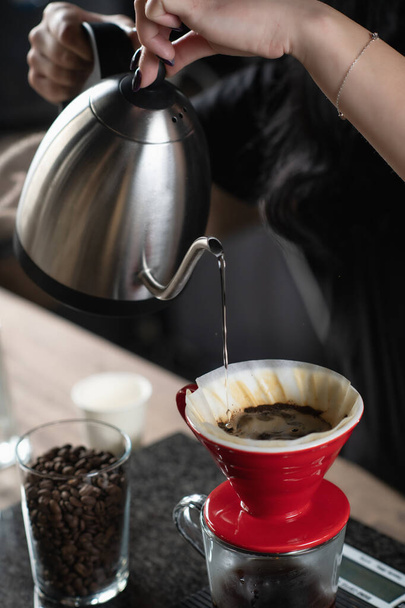 Γυναίκα barista ρίχνει ζεστό νερό από ένα βραστήρα σε κόκκους καφέ, δημιουργώντας ένα πλούσιο ρόφημα σε ένα προετοιμασμένο φίλτρο, σερβίρεται σε ένα φλιτζάνι καφέ. - Φωτογραφία, εικόνα